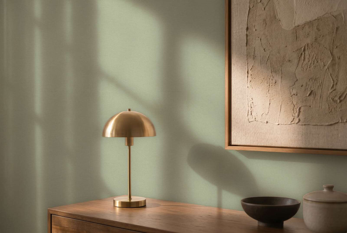 AS Creation Cosiness 792431 soft green textured wallpaper behind a wooden sideboard with a brass dome table lamp, framed textured artwork, and a ceramic bowl and lidded jar in warm light.