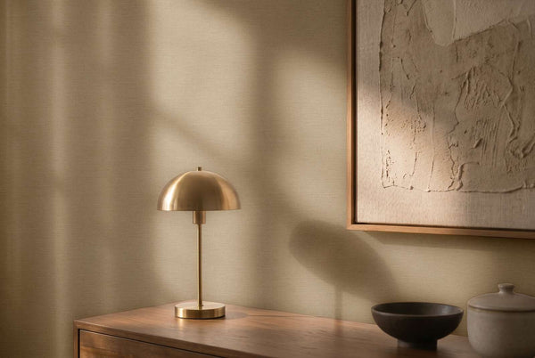 AS Creation Cosiness 792434 brown, Grey wallpaper with a subtle fine texture behind a brass dome table lamp, framed artwork, and a wooden console with two ceramic vessels.