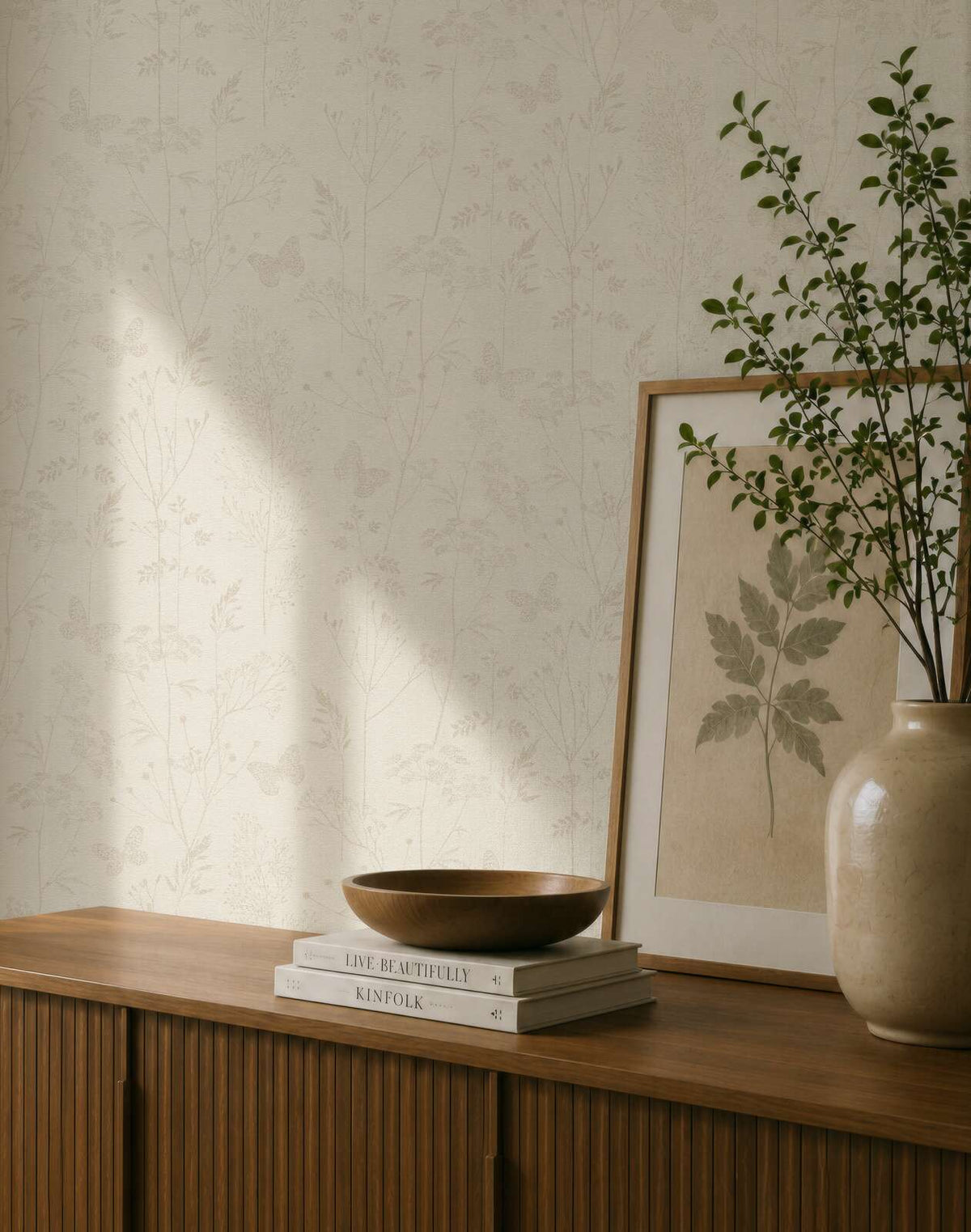 AS Creation Cosiness 792831 beige and brown wallpaper with a delicate botanical and butterfly pattern behind a wooden cabinet with a framed leaf print and a ceramic vase with branches.