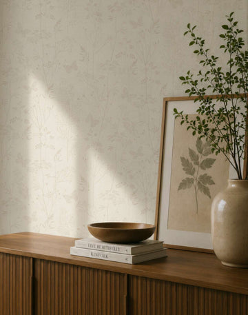 AS Creation Cosiness 792831 beige and brown wallpaper with a delicate botanical and butterfly pattern behind a wooden cabinet with a framed leaf print and a ceramic vase with branches.