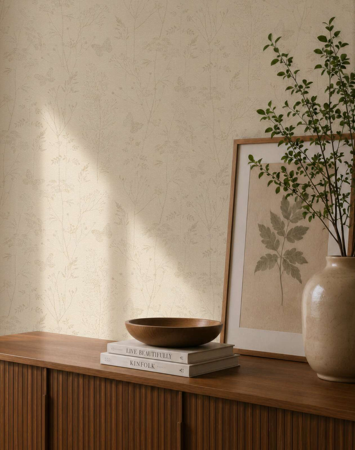 AS Creation Cosiness 792832 styled detail of Beige, Brown botanical wallpaper with delicate grasses and butterfly outlines behind a wooden sideboard with a ceramic vase and a framed leaf print.