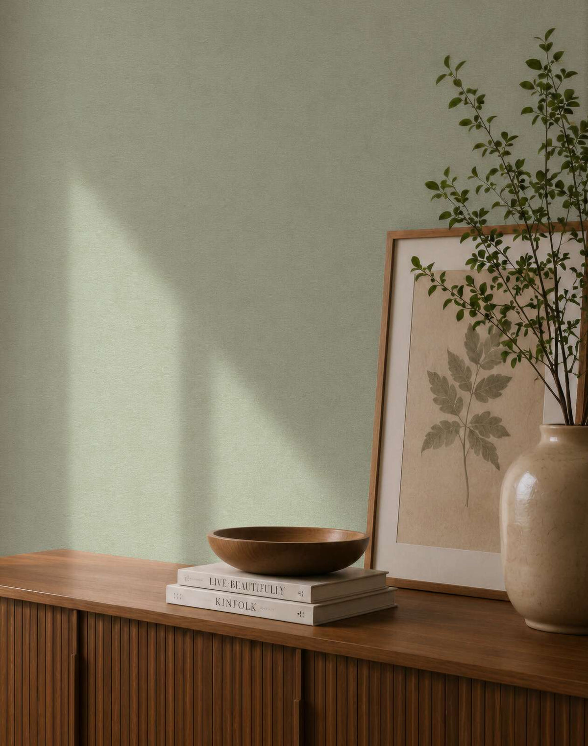 AS Creation Cosiness 792842 green wallpaper behind a wooden sideboard with a framed leaf print, a ceramic vase with branches, and a wooden bowl on stacked books.