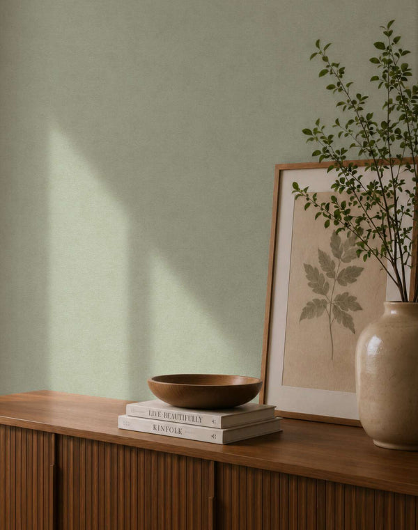AS Creation Cosiness 792842 green wallpaper behind a wooden sideboard with a framed leaf print, a ceramic vase with branches, and a wooden bowl on stacked books.