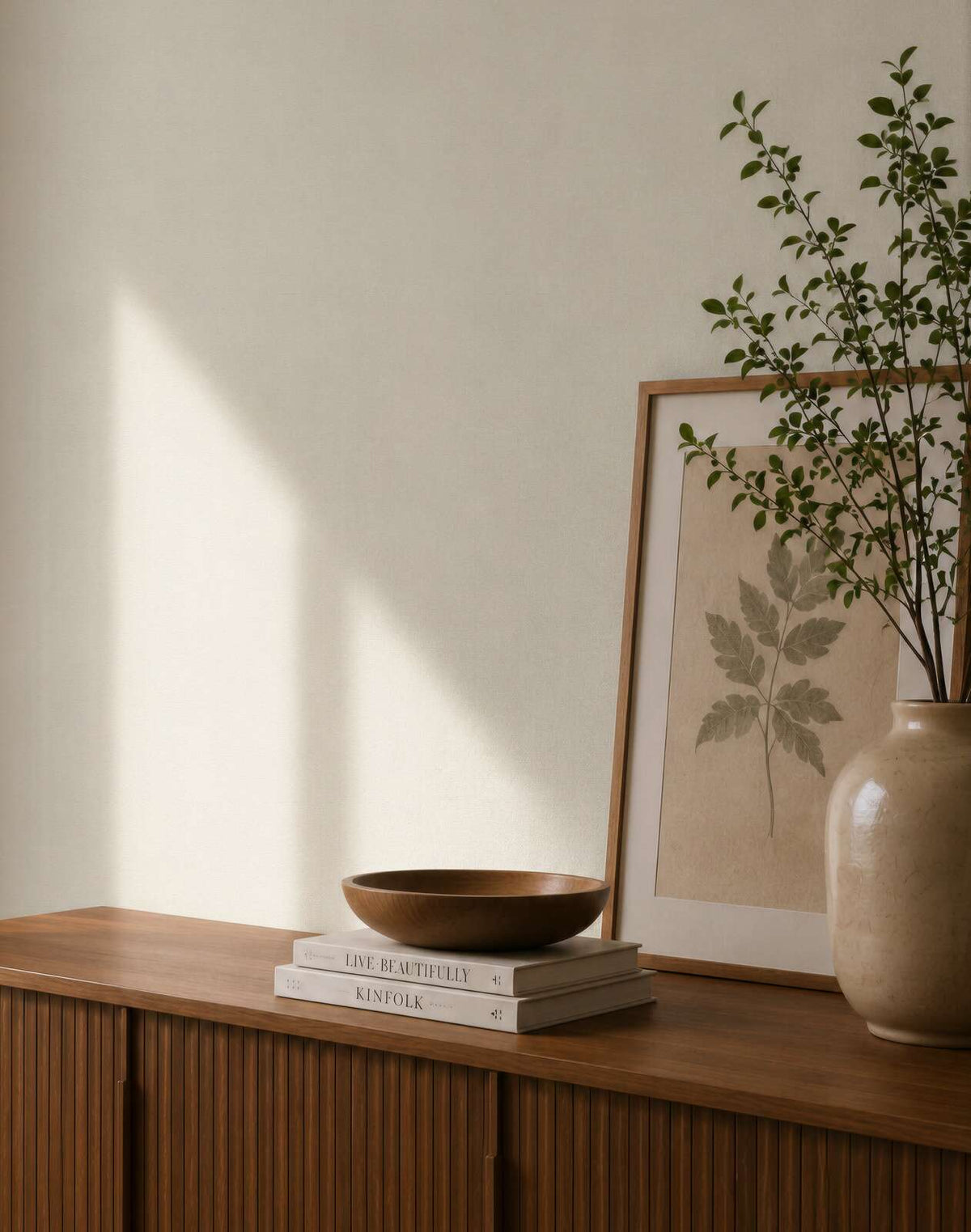 AS Creation Cosiness 792844 styled detail of Cream wallpaper behind a wooden cabinet with a framed leaf print, a ceramic vase with branches, and a wooden bowl on stacked books.