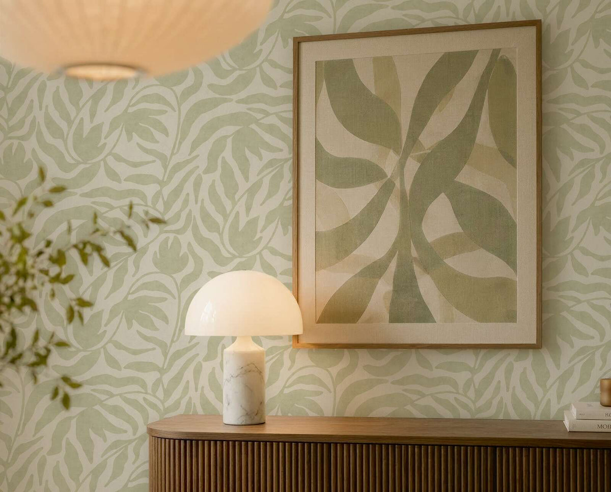 AS Creation Cosiness 793142 green botanical leaf motif wallpaper behind a wood console with a dome table lamp and framed art.
