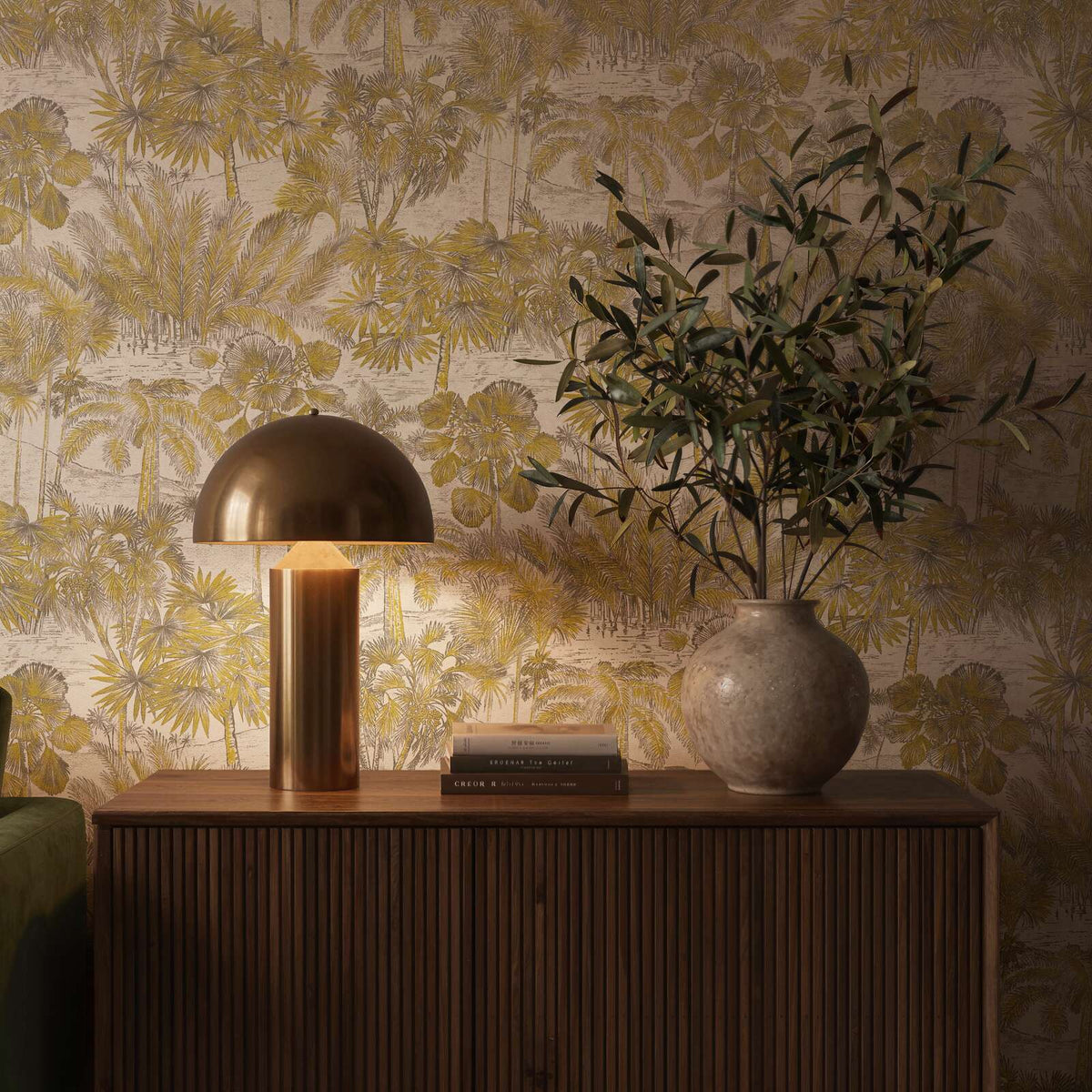AS Creation Cosiness 793171 scenic palm tree wallpaper in Beige, Brown, Metallic behind a wood sideboard with a dome lamp, books, and a round vase with branches.