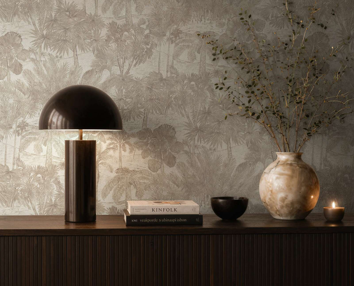 AS Creation Cosiness 793172 styled detail of beige and brown tropical palm wallpaper with metallic accents behind a console holding a dome table lamp, books, a bowl, a vase with branches, and a small candle.