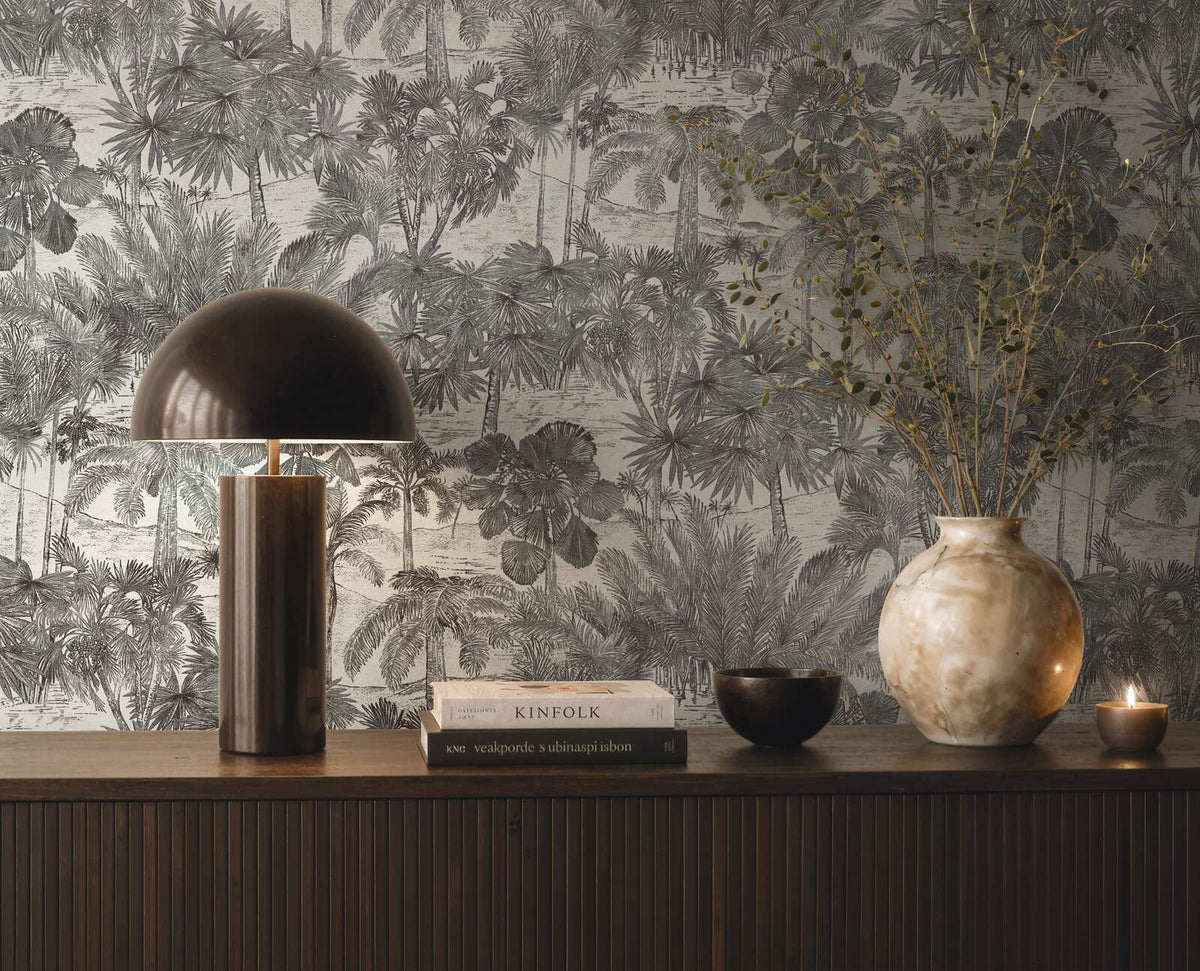 AS Creation Cosiness 793173 grey, Metallic tropical palm illustration wallpaper behind a console with a dome lamp, books, a large vase with branches, a small bowl, and a lit candle.