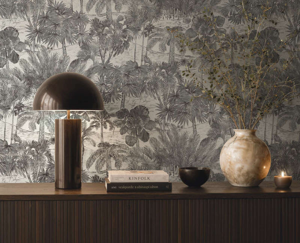 AS Creation Cosiness 793173 grey, Metallic tropical palm illustration wallpaper behind a console with a dome lamp, books, a large vase with branches, a small bowl, and a lit candle.