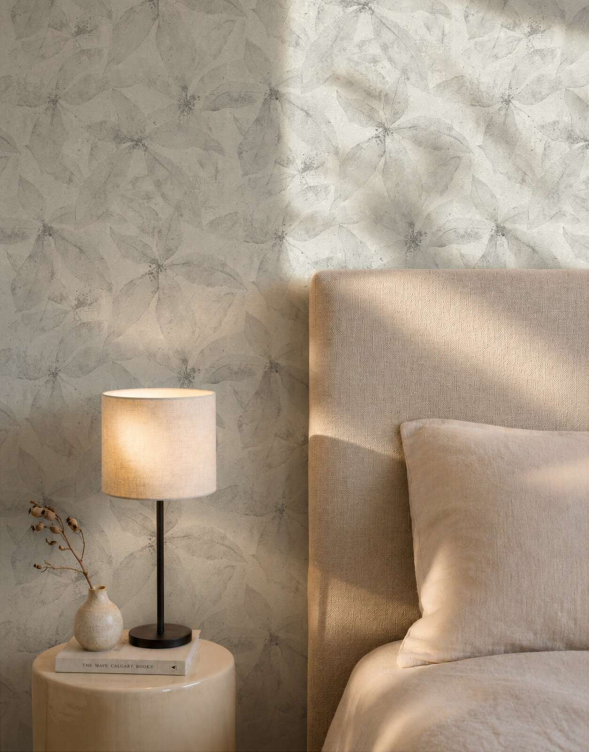 AS Creation Cosiness 793203 bedroom scene with Grey wallpaper featuring a delicate overlapping botanical motif; a bed with an upholstered headboard and a table lamp on a round side table are visible.