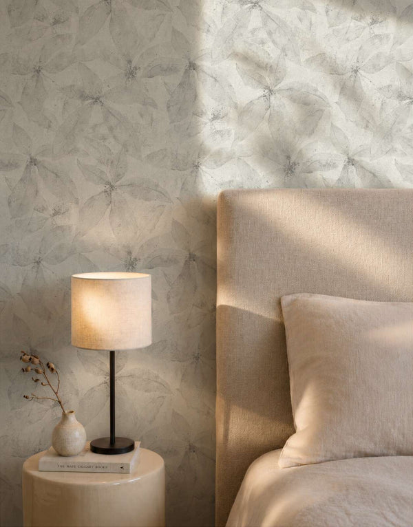 AS Creation Cosiness 793203 bedroom scene with Grey wallpaper featuring a delicate overlapping botanical motif; a bed with an upholstered headboard and a table lamp on a round side table are visible.