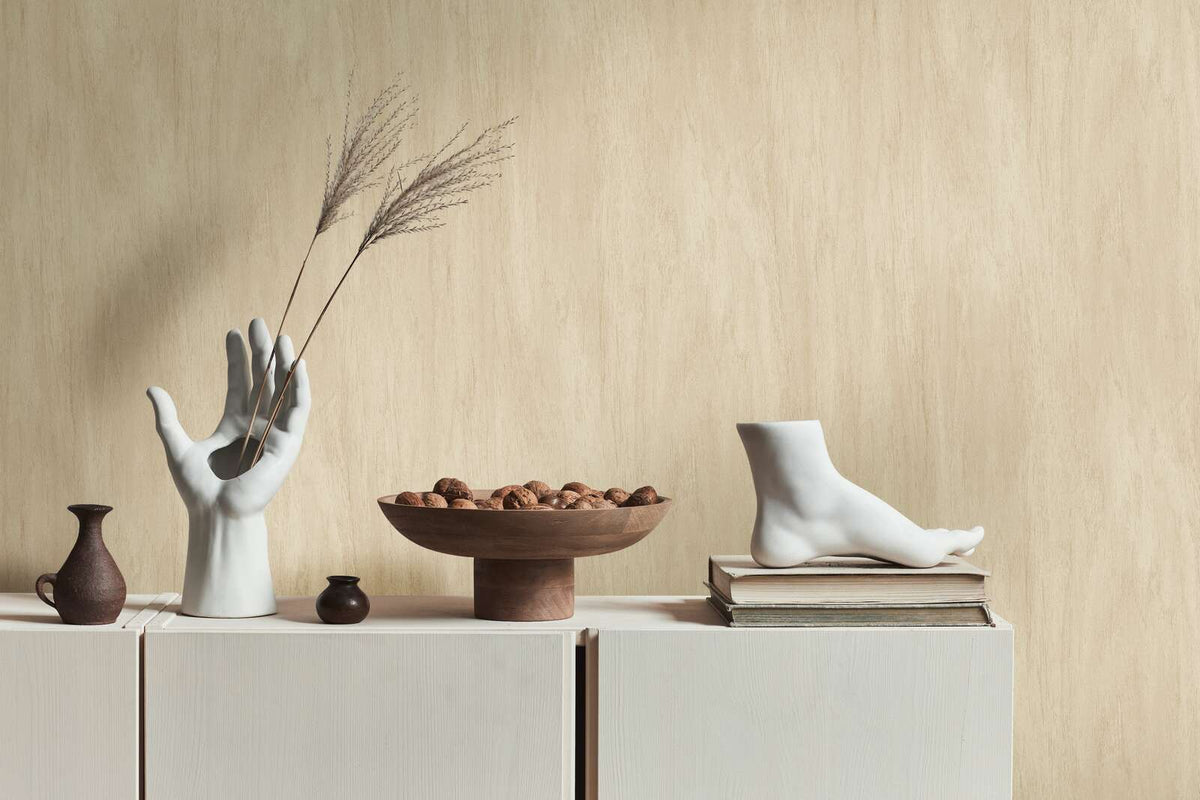 AS Creation French Affair 398011 beige, Cream wallpaper with soft vertical tonal variation behind a light cabinet styled with a white hand sculpture holding dried stems, a wooden bowl of nuts, a small jug and pot, and a white foot sculpture on books.