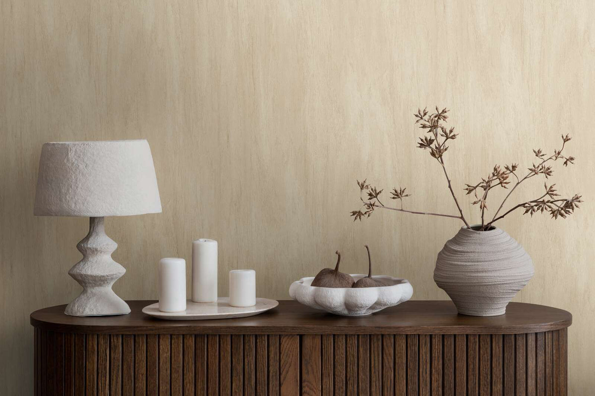 AS Creation French Affair 398011 beige and Cream wallpaper with soft vertical tonal striations behind a wood console styled with a sculptural lamp, candles, a bowl with decorative pears, and a vase with branches.