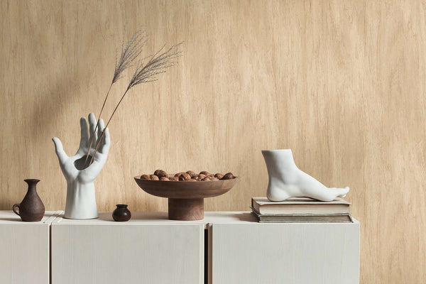 AS Creation French Affair 398012 beige wallpaper with a subtle vertical strie effect behind a low cabinet styled with a hand sculpture holding stems, a wooden bowl of nuts, and a foot sculpture on stacked books.