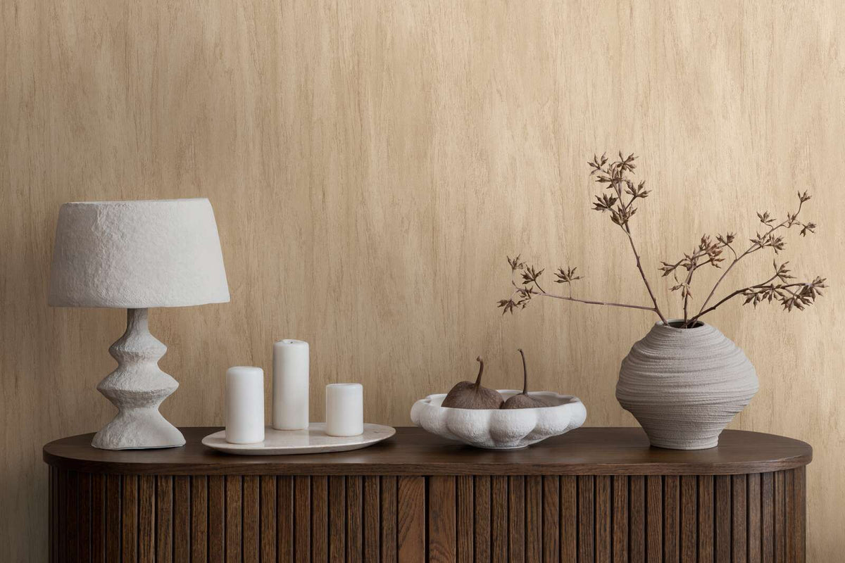 AS Creation French Affair 398012 beige wallpaper with a soft vertical strie behind a wood sideboard styled with a sculptural lamp, pillar candles on a tray, a bowl, and a ceramic vase with branches.