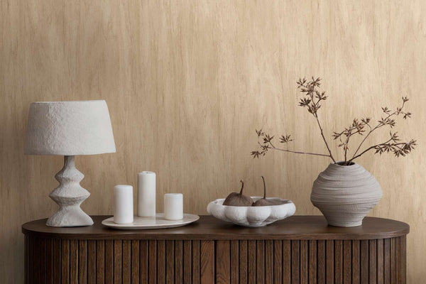 AS Creation French Affair 398012 beige wallpaper with a soft vertical strie behind a wood sideboard styled with a sculptural lamp, pillar candles on a tray, a bowl, and a ceramic vase with branches.