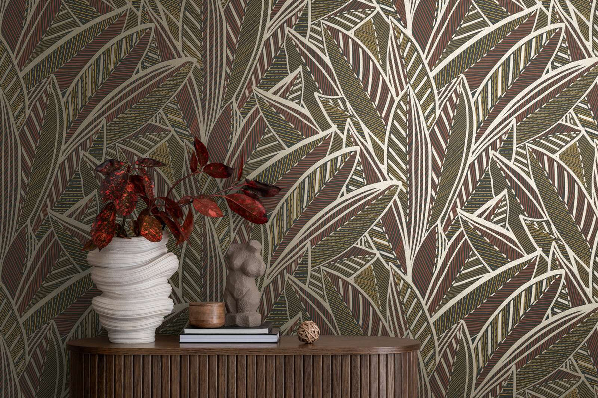 AS Creation French Affair 399331 leaf pattern wallpaper in Beige, Black, Brown, Gold, Red, Yellow behind a curved wood sideboard with a sculptural white vase holding red foliage and a small figure sculpture on books.
