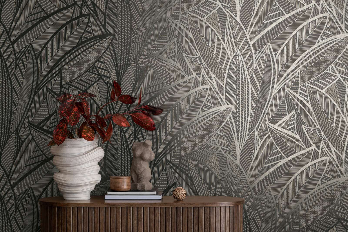 AS Creation French Affair 399334 styled detail of Black, Gold, Metallic leaf pattern wallpaper behind a wood console with a sculptural vase, a small figure sculpture, and books.