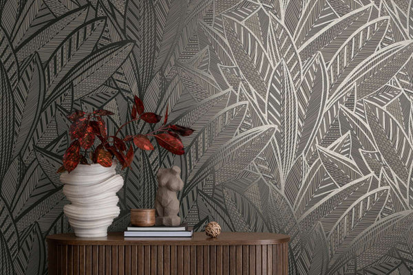 AS Creation French Affair 399334 styled detail of Black, Gold, Metallic leaf pattern wallpaper behind a wood console with a sculptural vase, a small figure sculpture, and books.