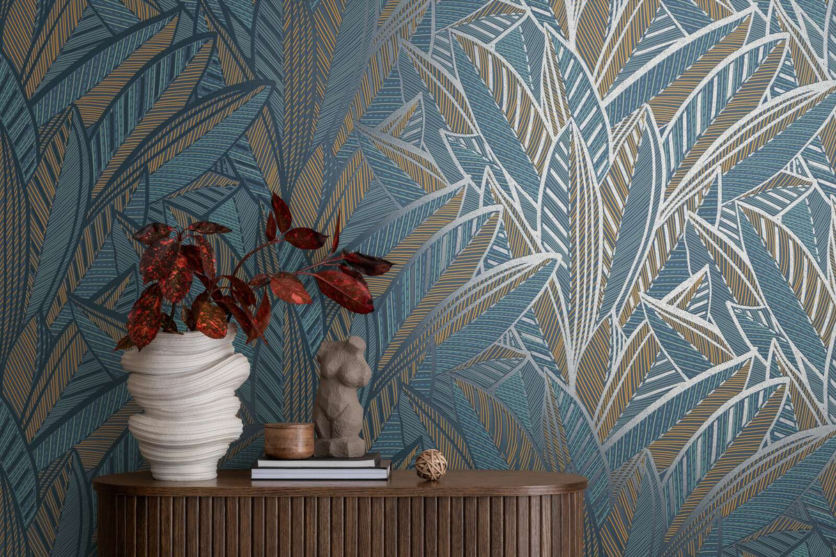 AS Creation French Affair 399335 graphic leaf wallpaper in Blue, Green, Turquoise, and Yellow behind a console with a sculptural vase holding red foliage, a small torso sculpture, and books.