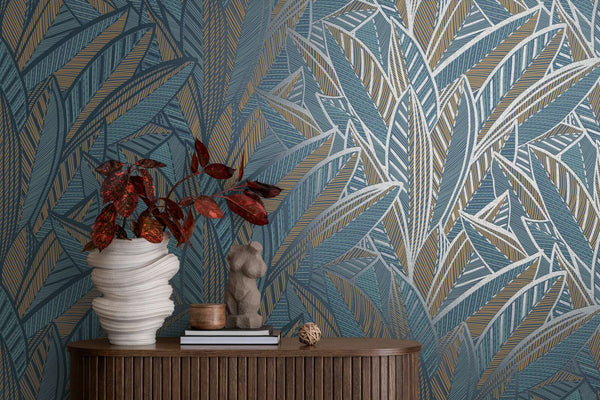 AS Creation French Affair 399335 graphic leaf wallpaper in Blue, Green, Turquoise, and Yellow behind a console with a sculptural vase holding red foliage, a small torso sculpture, and books.