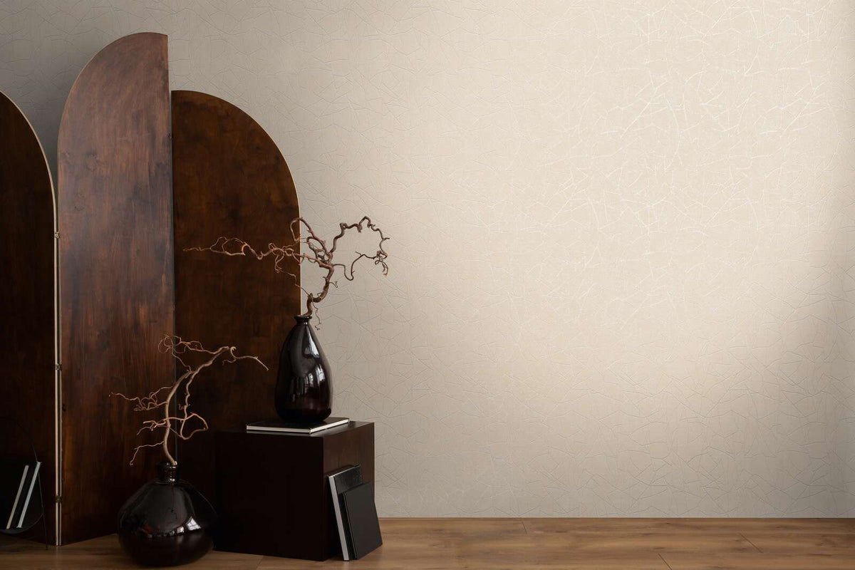 AS Creation French Affair 399342 beige and cream wallpaper with a subtle white abstract line pattern, styled with a dark wood screen, two dark vases with branches, a small pedestal, and books on a wood floor.