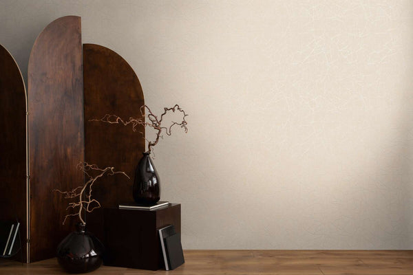 AS Creation French Affair 399342 beige and cream wallpaper with a subtle white abstract line pattern, styled with a dark wood screen, two dark vases with branches, a small pedestal, and books on a wood floor.