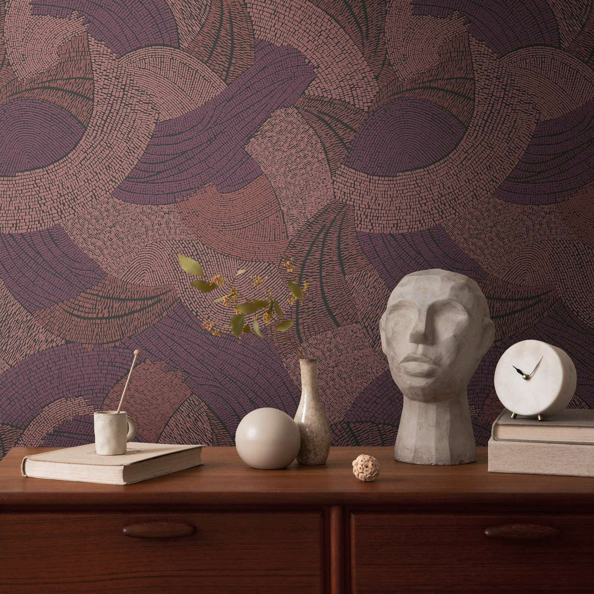 AS Creation French Affair 399352 red abstract wallpaper with overlapping curved lines in sweeping arcs behind a wood sideboard styled with a sculptural head, a round clock, and a small vase.