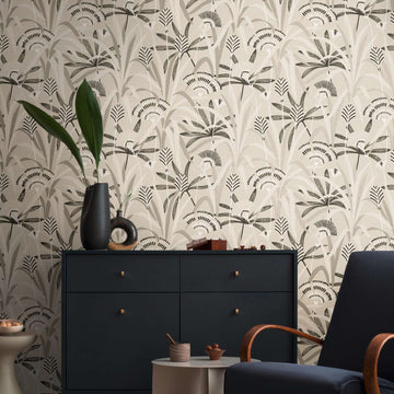 AS Creation French Affair 399361 living room with botanical wallpaper in Beige, Black, Grey, Taupe, and White, shown behind a dark sideboard and an armchair.