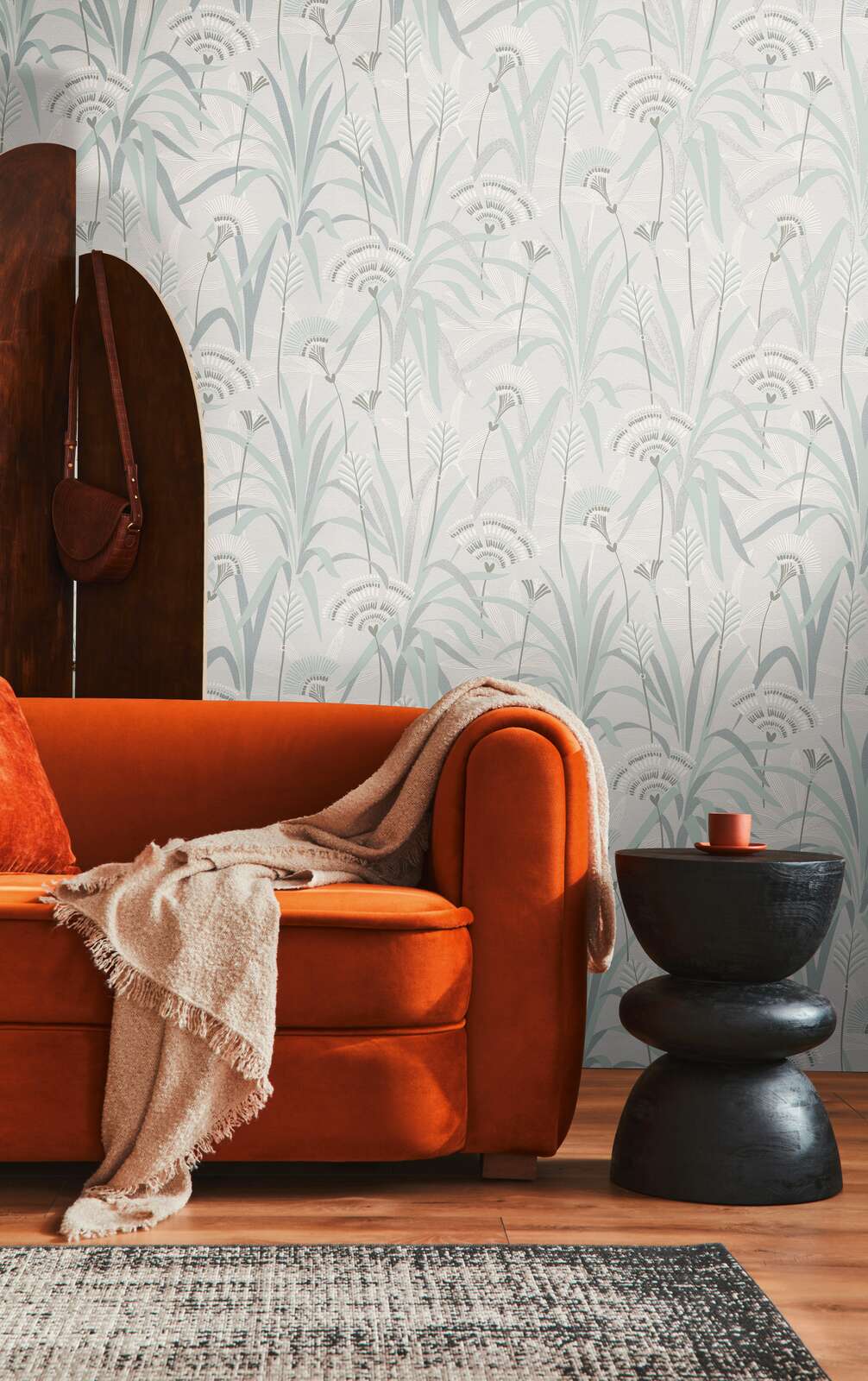 AS Creation French Affair 399363 living room featuring a botanical wallpaper in green, grey, and turquoise on white, shown behind an orange sofa and a black side table.