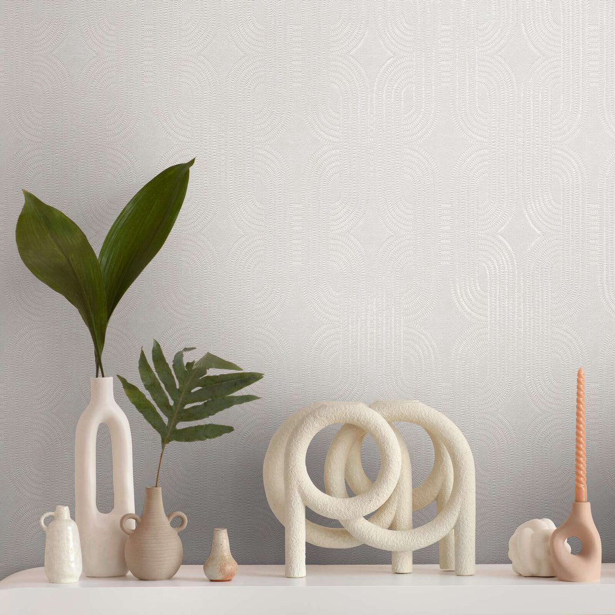 AS Creation French Affair 399372 grey and White geometric wallpaper with repeating arches behind a styled tabletop holding ceramic vases with green leaves, a looped sculpture, and a twisted taper candle.