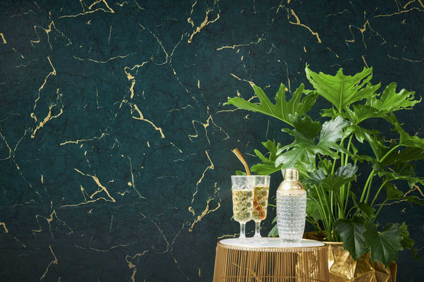 AS Creation Metropolitan Stories Ii 378555 green wallpaper with metallic marbled veining behind a small round side table holding two drinks and a cocktail shaker next to a leafy plant.