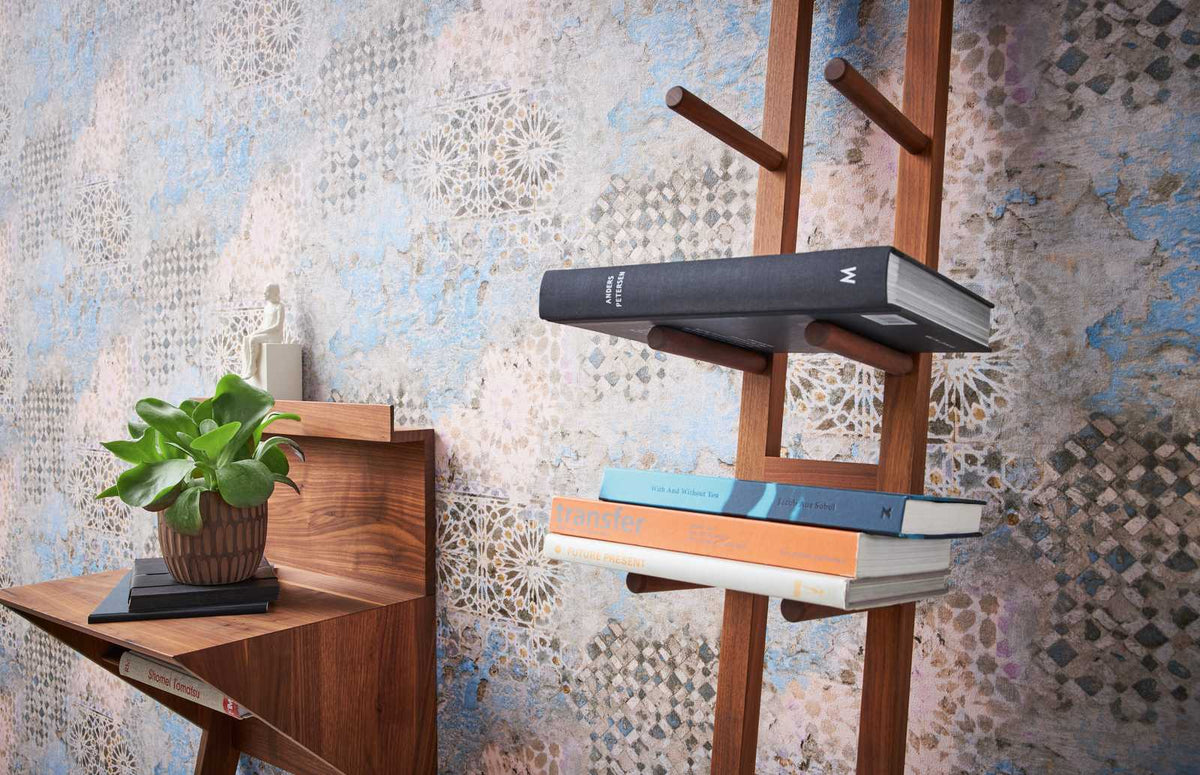 AS Creation Metropolitan Stories Ii 378581 beige, Blue, Brown wallpaper with a distressed ornamental pattern of rosettes and diamond lattice behind a wooden ladder shelf of books and a small console with a potted plant.