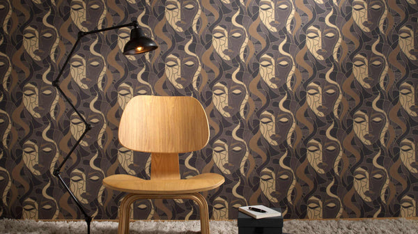 Erismann Elle Decoration abstract face wallpaper in brown and gold tones styled with a wooden chair and modern floor lamp in an interior setting