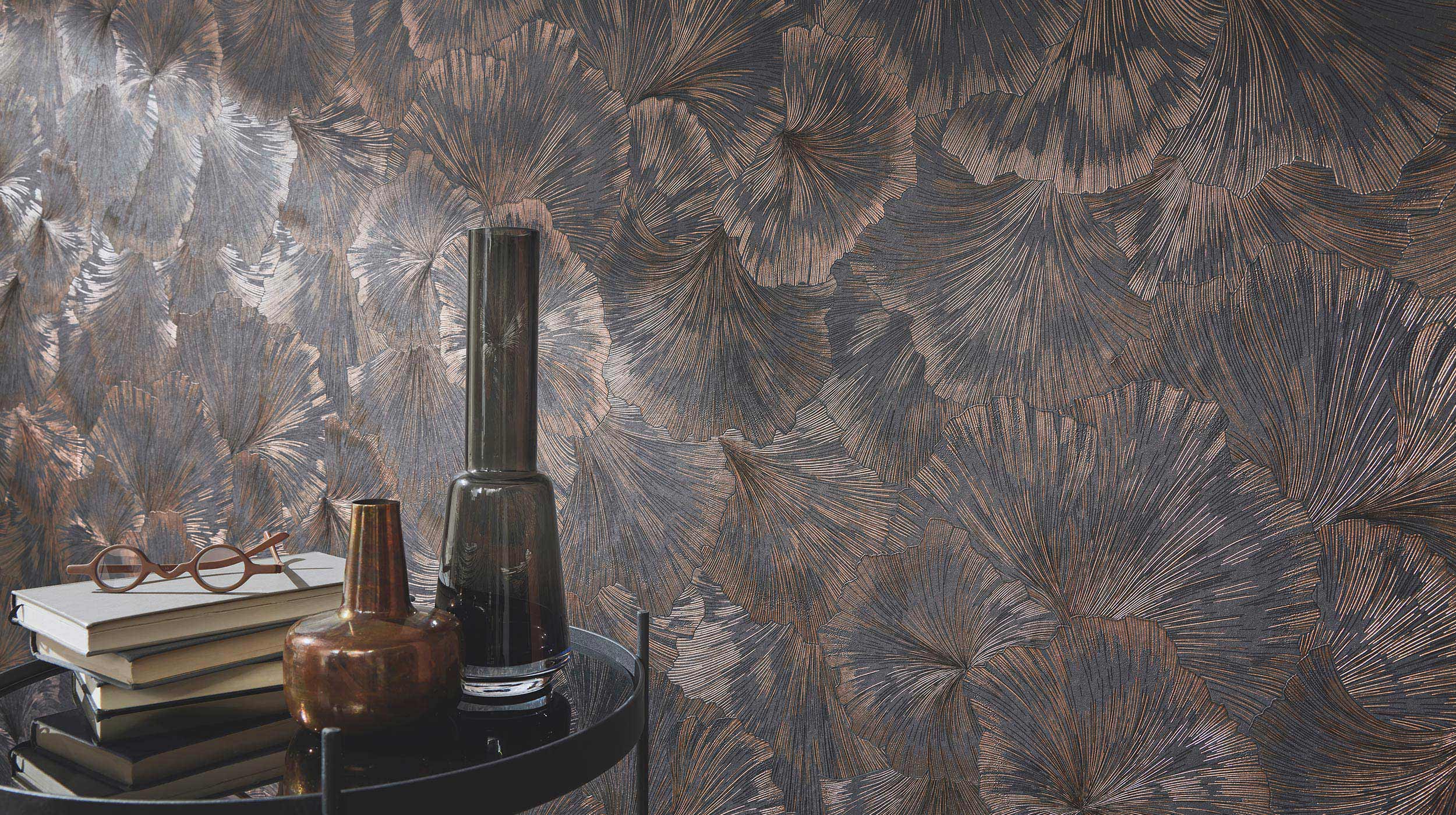 Erismann Fashion For Walls 4 10373-47 wallpaper lounge feature wall with dark metallic botanical pattern behind a round side table holding stacked books, glasses, and vases for a moody, sophisticated feel.
