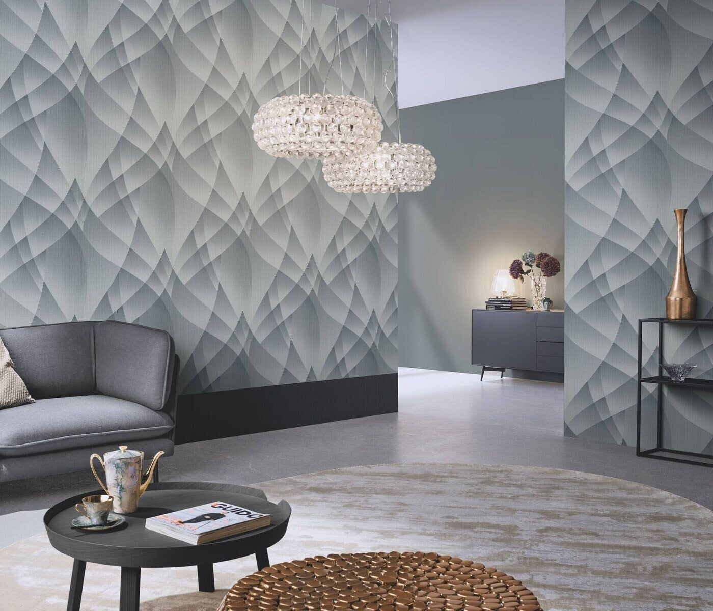 Erismann Fashion For Walls 4 10374-29 wallpaper in a modern lounge with a geometric feature wall, curved sofa, round coffee table with tea set and magazine, bubble pendant lights, sideboard with lamp and vases, and a calm contemporary feel.