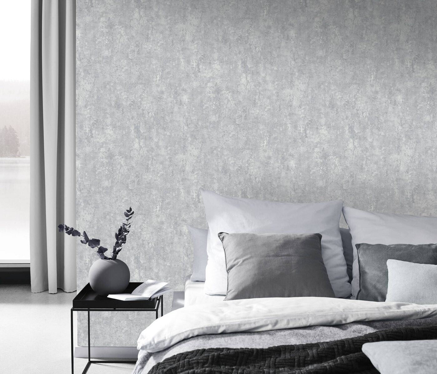 Erismann Fashion For Walls 4 10375-31 wallpaper bedroom feature wall behind a neatly made bed with layered grey pillows, slim black side table with round vase and book, curtains, calm minimalist mood.