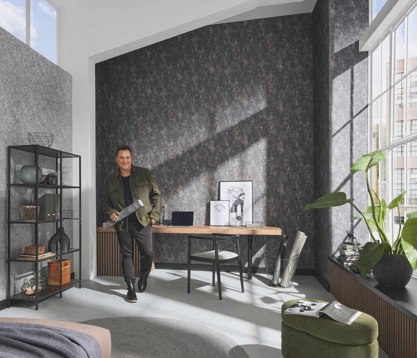 Erismann Fashion For Walls 4 10375-47 wallpaper lounge with a dark patterned feature wall behind a wooden desk, framed art and laptop, black metal shelving, large window with potted plant and green ottoman, bright modern vibe.