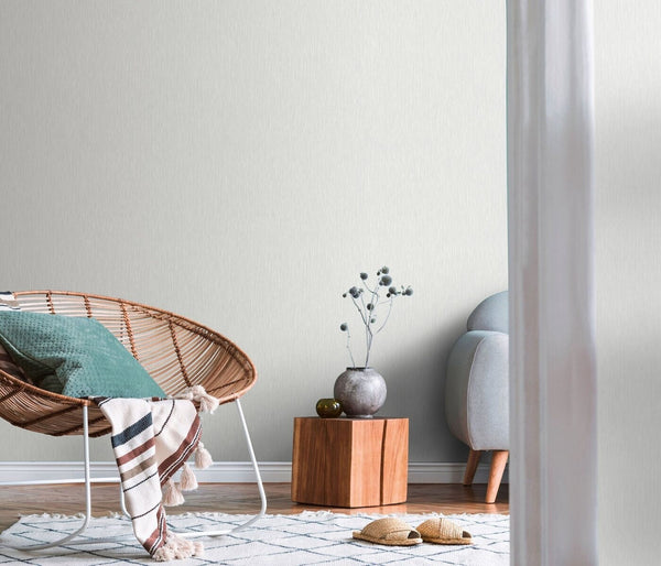 Erismann Fashion For Walls 4 10376-31 wallpaper lounge feature wall behind rattan lounge chair with cushion and striped throw, wood block side table with vase and stems, partial soft armchair, patterned rug and slippers, calm minimalist mood.