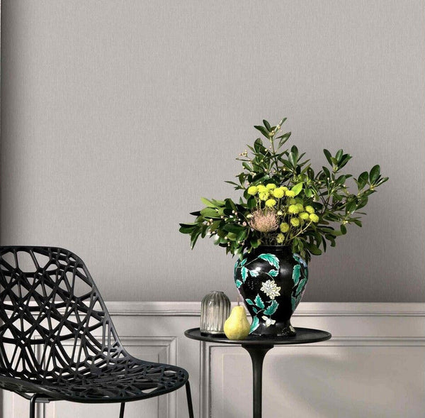 Erismann Fashion For Walls 4 10376-38 wallpaper in a lounge with a soft grey feature wall above paneling, black lattice chair, round side table, and an ornate vase of greenery with a pear, calm modern mood.
