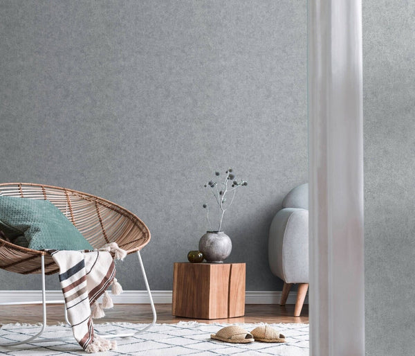 Erismann Fashion For Walls 4 10377-29 wallpaper lounge feature wall with textured grey finish, rattan chair with cushion and striped throw, wood block side table with vase, rug and slippers, calm minimalist mood.