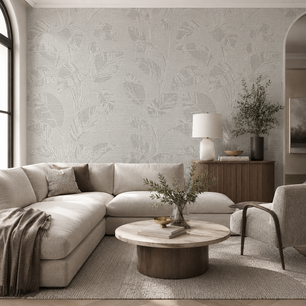 Erismann Opus 10459-24 soft grey botanical leaf wallpaper in a bright modern living room with a cream sectional sofa, round marble coffee table, textured rug, wood cabinet, and neutral organic decor.