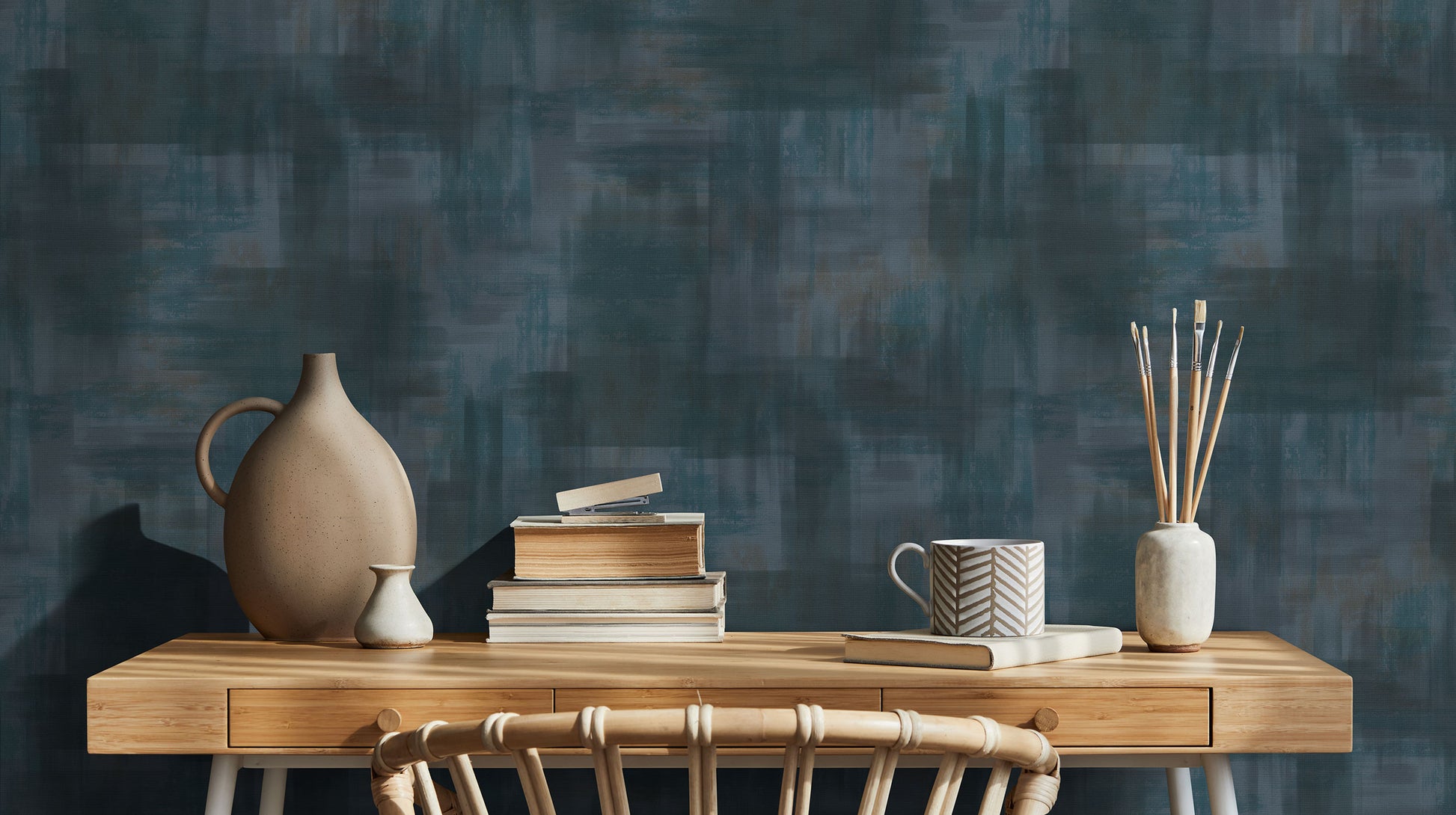 Erismann Elysium abstract wallpaper in deep teal and blue tones with layered painterly textures, styled with wooden furniture, books, ceramics, and artist brushes.