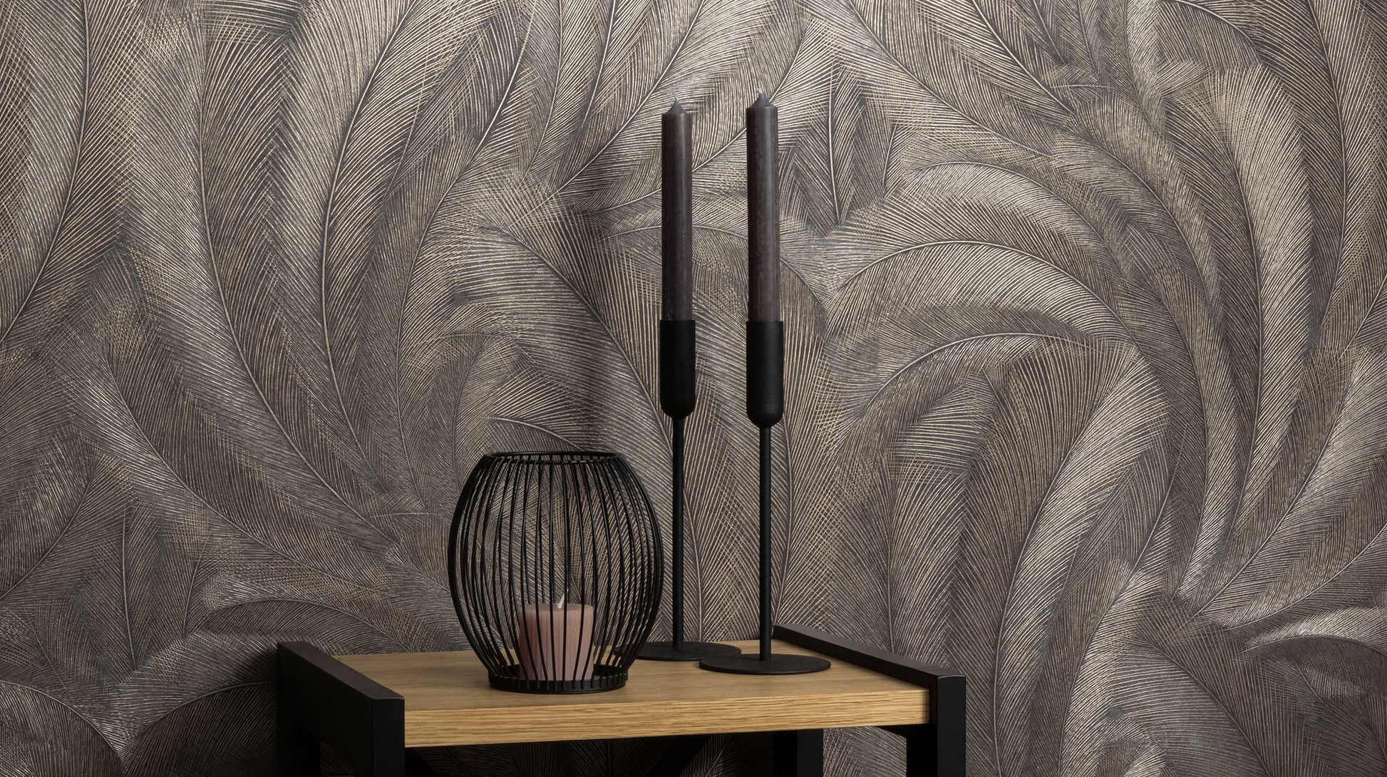 Erismann Elysium botanical feather wallpaper in deep charcoal and metallic taupe tones, styled as a feature wall behind a console table with decorative candles and accents, creating a moody and sophisticated atmosphere with rich texture and subtle shimmer.