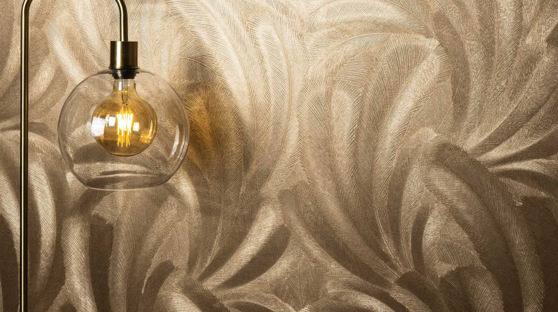 Erismann Elysium botanical feather wallpaper in warm gold tones, styled as a feature wall with a glass pendant light, creating a rich and luxurious ambiance that highlights texture and enhances the glow of soft lighting in a modern interior.