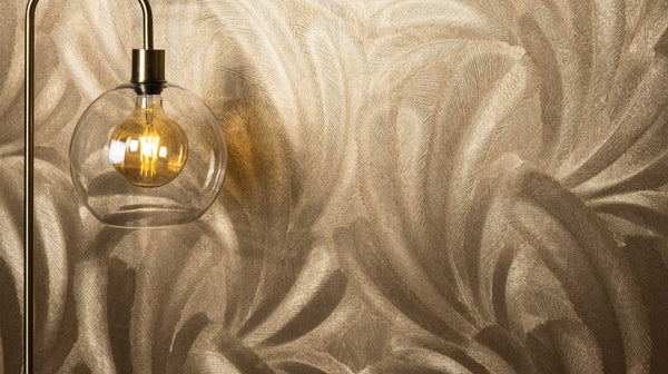 Erismann Elysium botanical feather wallpaper in warm gold tones, styled as a feature wall with a glass pendant light, creating a rich and luxurious ambiance that highlights texture and enhances the glow of soft lighting in a modern interior.