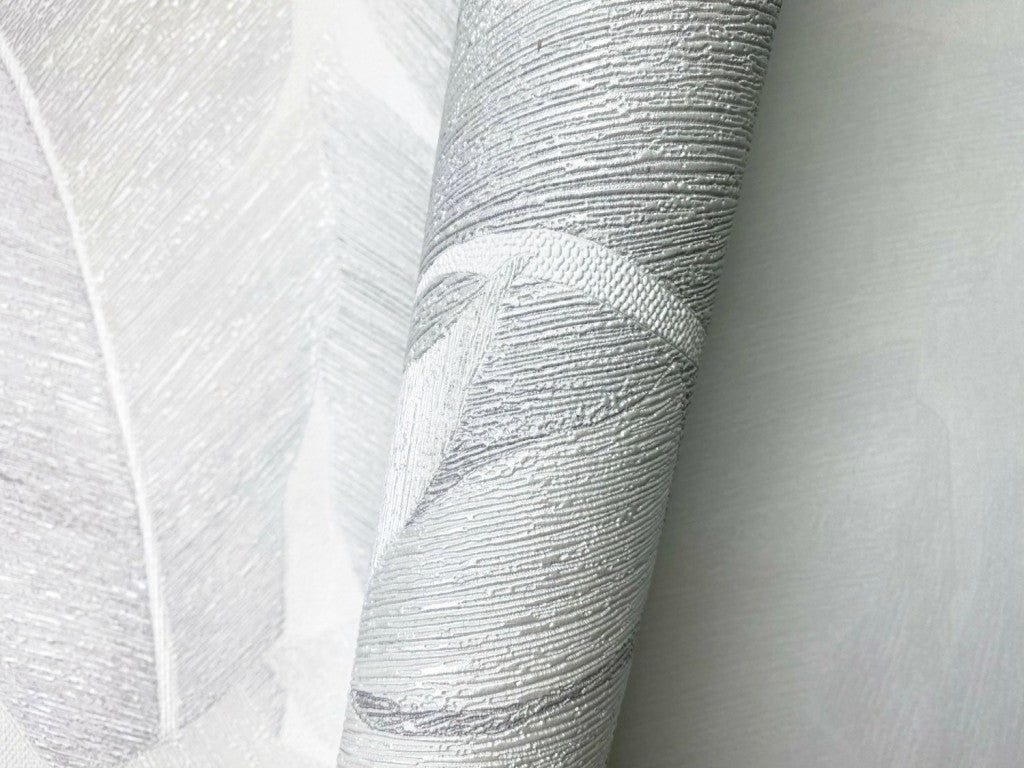 Marburg Avalon 31623 Close-up of a textured wallpaper roll with an embossed silver and light gray leaf pattern resting on the same design, showing raised metallic detailing.