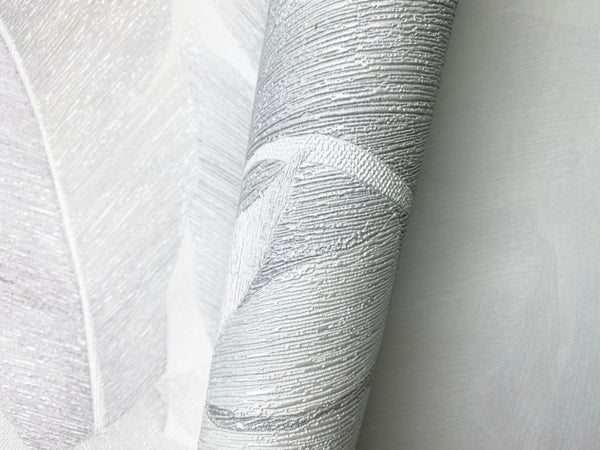 Marburg Avalon 31623 Close-up of a textured wallpaper roll with an embossed silver and light gray leaf pattern resting on the same design, showing raised metallic detailing.