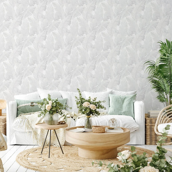 Marburg Avalon 31623 Light gray leaf-pattern wallpaper behind a white sofa with pale green pillows, a round wooden coffee table, woven rug, rattan accents, and indoor plants with flower vases.