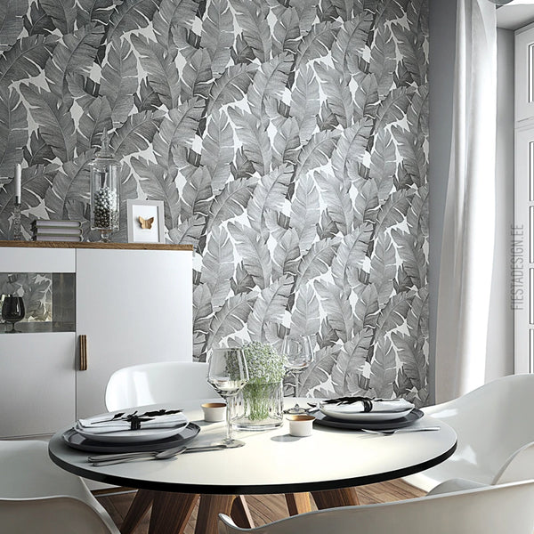 Marburg Avalon 31624 Grayscale tropical banana leaf wallpaper on a dining room wall, shown with a round table set with plates and glasses, white chairs, a sideboard, and a window with curtains.